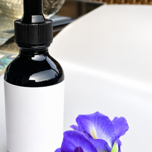 A dark glass dropper bottle containing Blue Lotus liquid ceremonial extract placed next to vibrant blue botanical flowers on a clean surface.